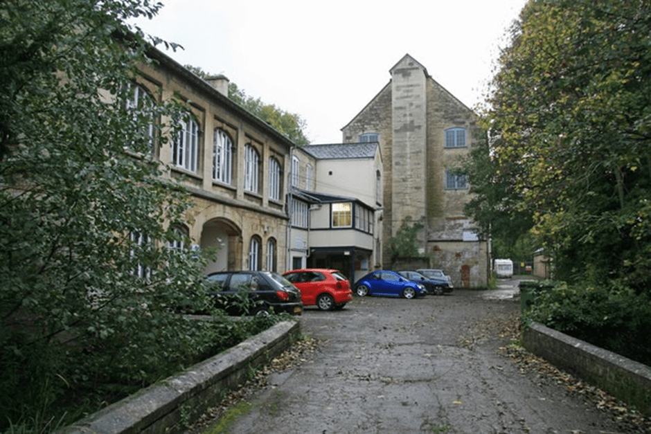 A photo over a small bridge of an old mill, with cars parked outside 