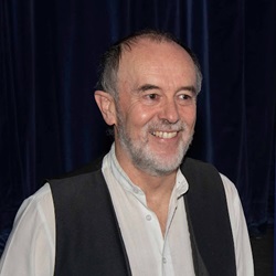 A man in a white shirt and a waistcoat looking out and left, smiling 