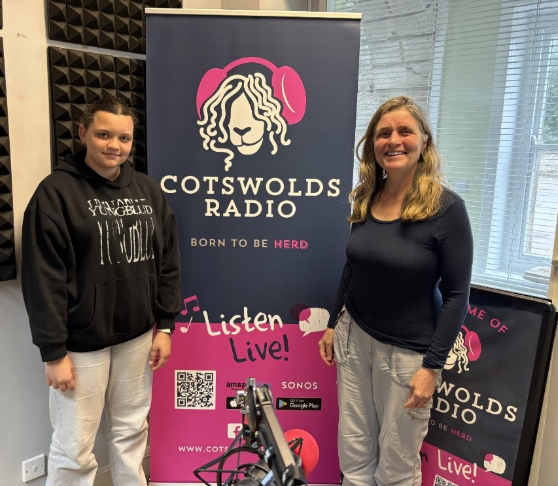Two people looking at the camera and standing in front of a banner for "Cotswolds Radio"